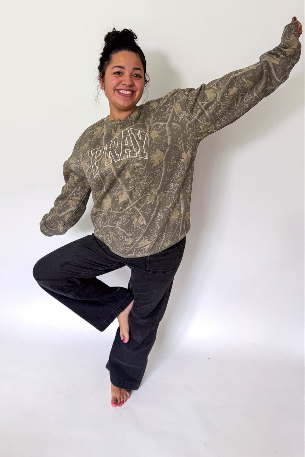 A woman posing in a vintage-style camouflage sweatshirt with 'PRAY' printed in arched block letters on the chest, paired with black pants, against a plain white studio background.