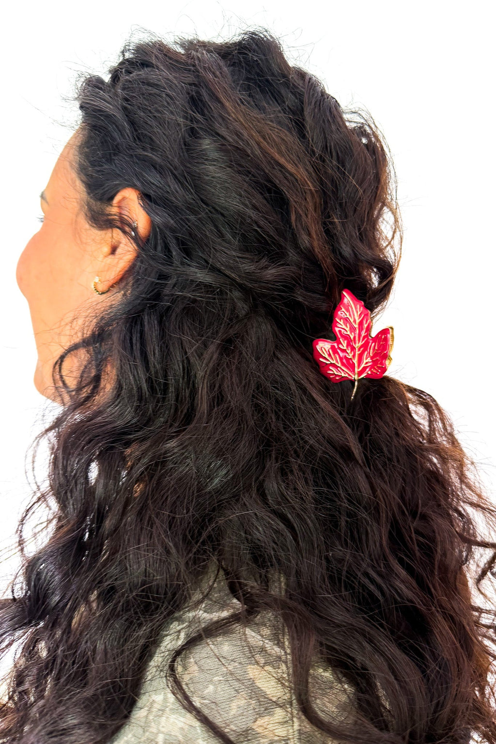 A close-up side profile of a woman with long, wavy dark brown hair wearing a red maple leaf hair clip, set against a plain white background.