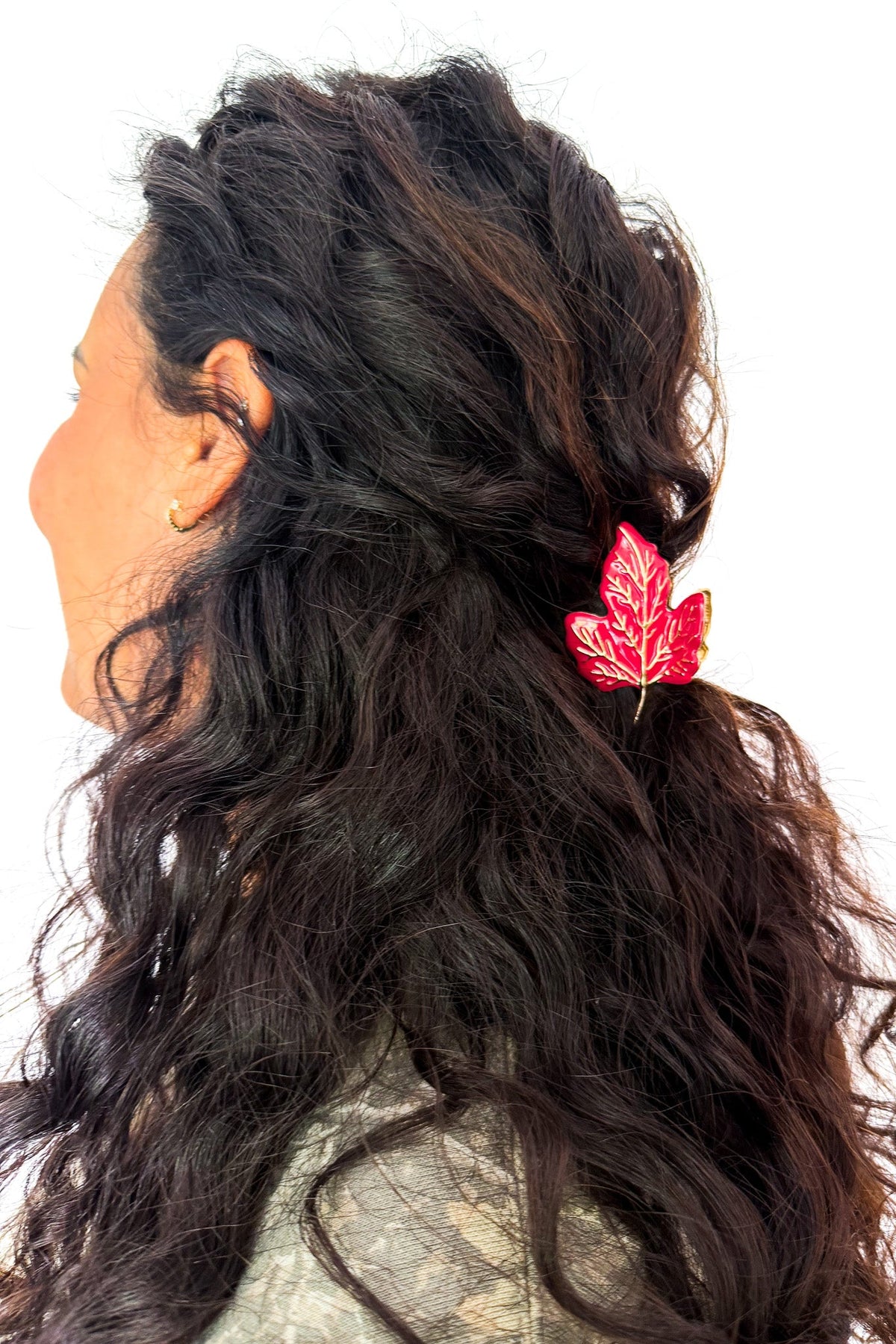 A close-up side profile of a woman with long, wavy dark brown hair wearing a red maple leaf hair clip, set against a plain white background.