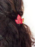 A close-up of a person's dark brown wavy hair featuring a red maple leaf hair clip with gold leaf detailing and a small gold butterfly accent, set against a plain white background.