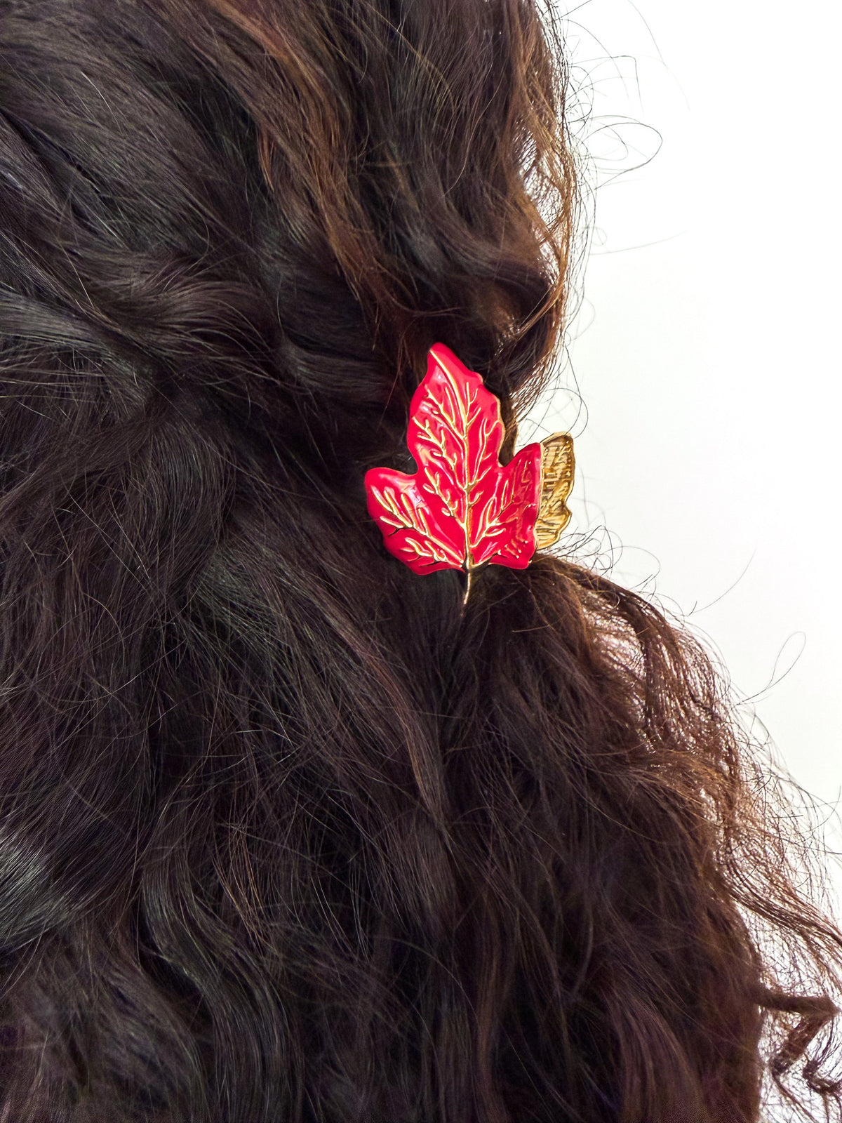 A close-up of a person's dark brown wavy hair featuring a red maple leaf hair clip with gold leaf detailing and a small gold butterfly accent, set against a plain white background.