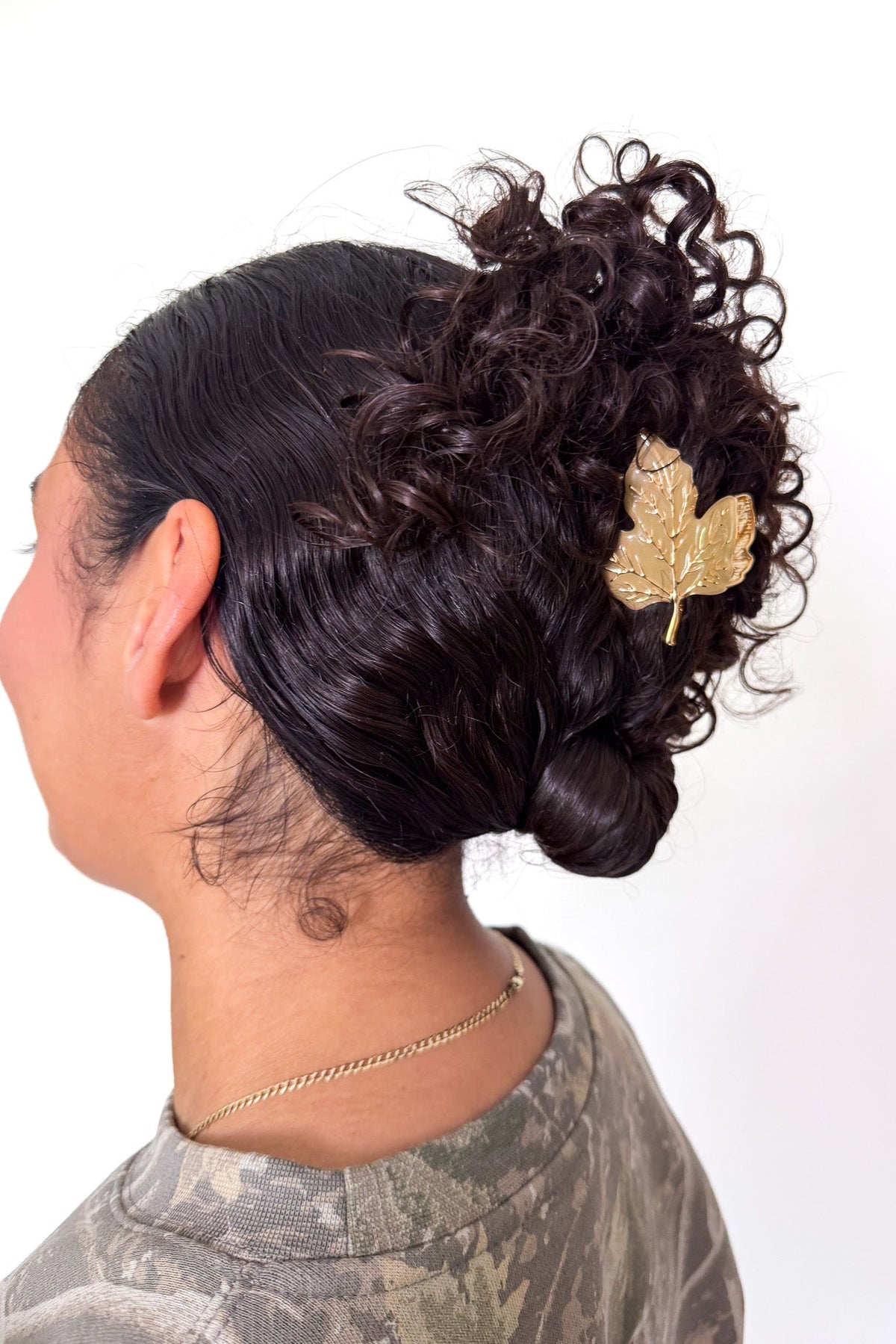 A person shown from the side and back wearing a dark curly bun hairstyle with a gold leaf-shaped hair accessory, against a plain white background.