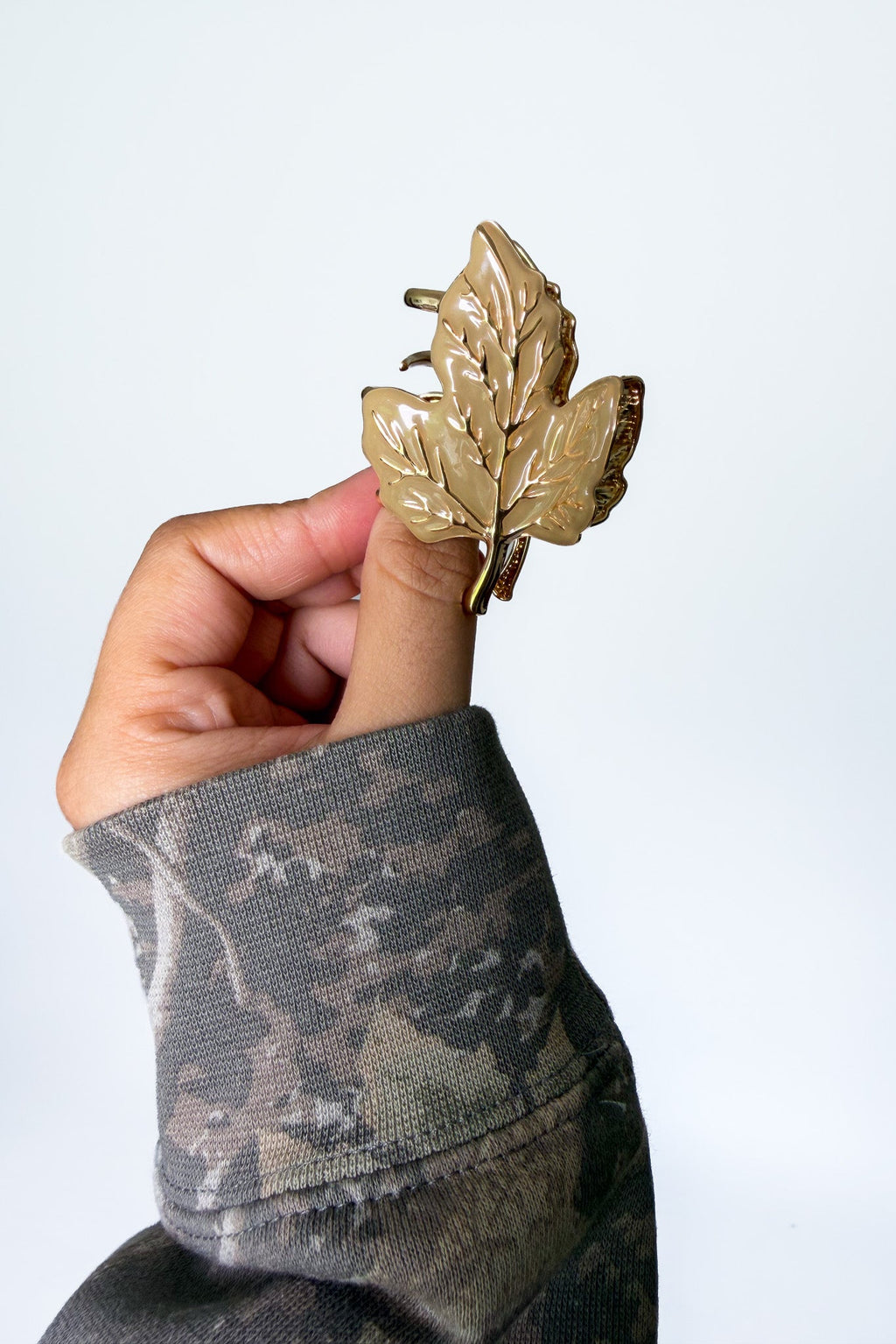 A hand wearing a camouflage sleeve holds a metallic gold leaf-shaped hair claw clip against a plain light gray background.