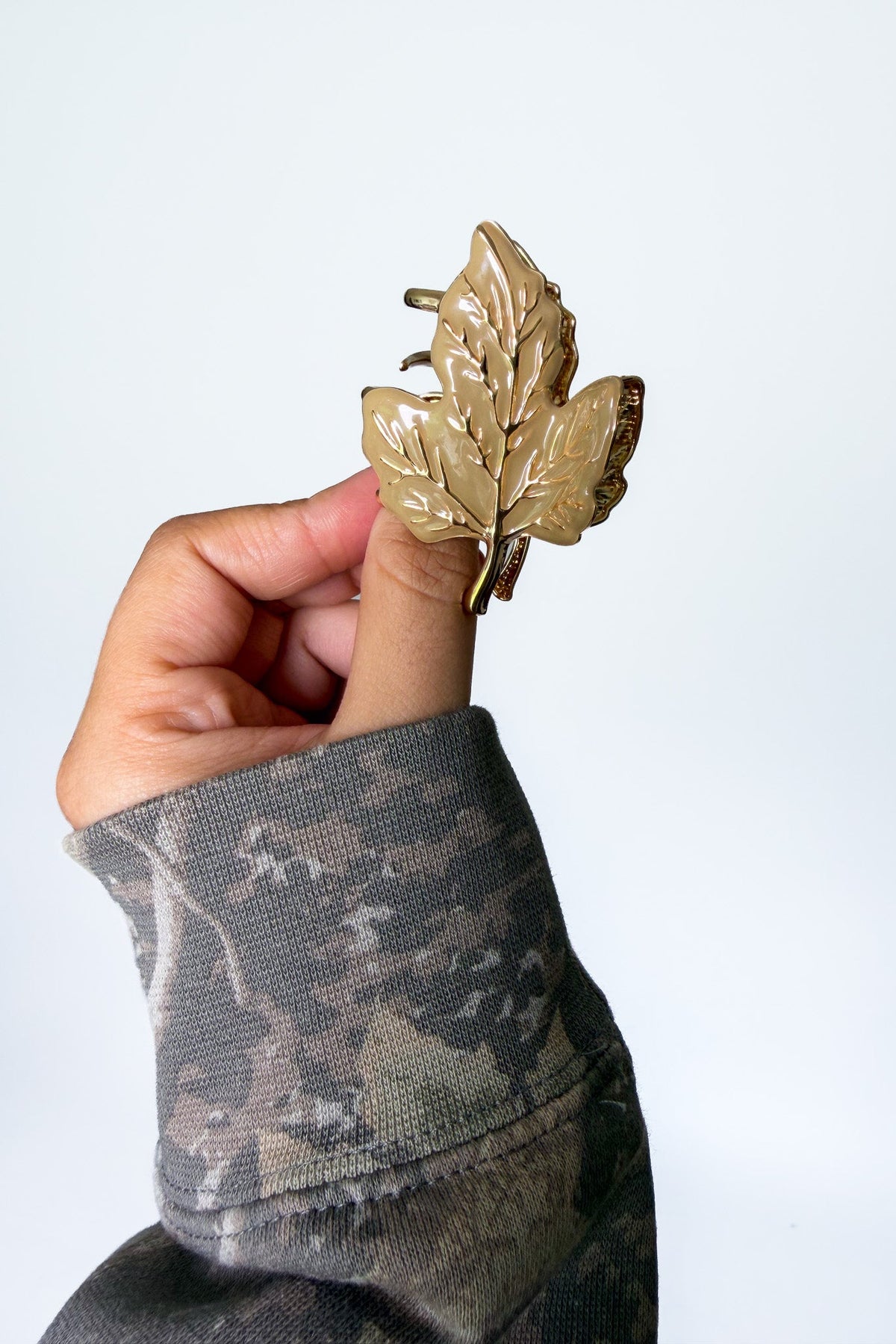 A hand wearing a camouflage sleeve holds a metallic gold leaf-shaped hair claw clip against a plain light gray background.