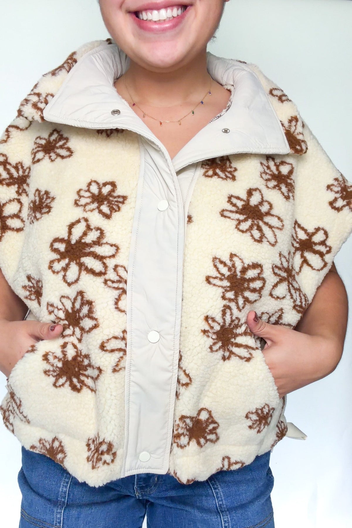 A person wearing a cream sherpa fleece zip-up vest with brown floral patterns, paired with blue jeans, against a plain white background.