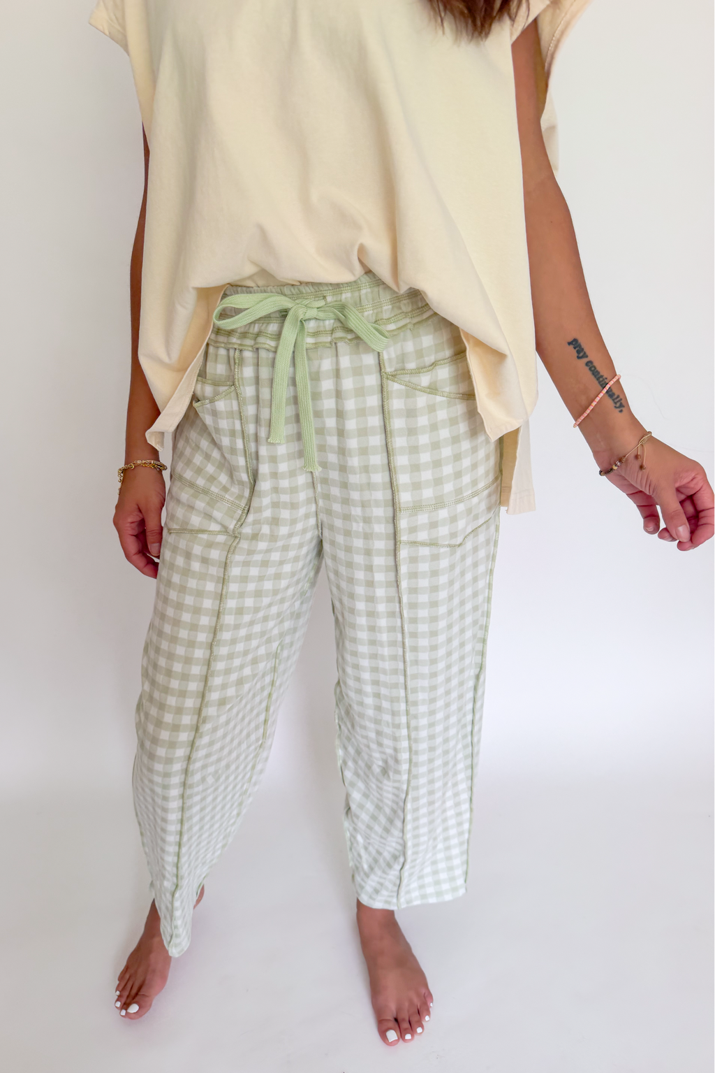 A person wearing light sage green and white gingham cropped pants with a matching sage green drawstring waist, styled with a cream-colored top against a plain white background.