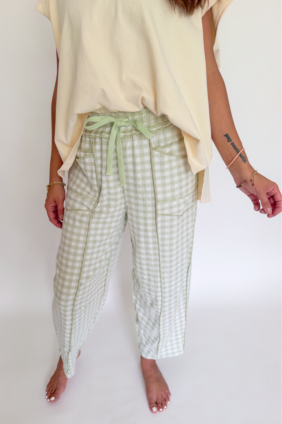 A person wearing light sage green and white gingham cropped pants with a matching sage green drawstring waist, styled with a cream-colored top against a plain white background.