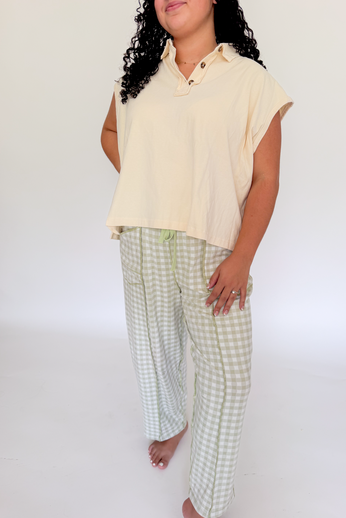 A woman modeling a two-piece set consisting of a light cream short-sleeve collared top and light green and white gingham pants, set against a plain white studio background.