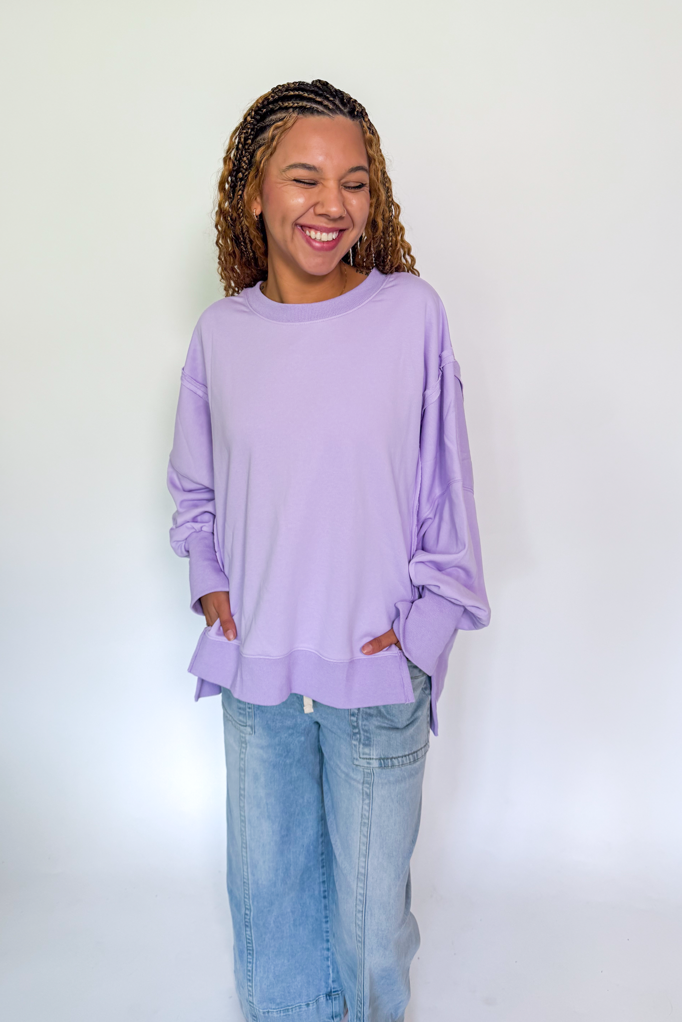 A woman with braided hair wearing a lavender long-sleeve top and light blue jeans, standing against a plain white studio background.