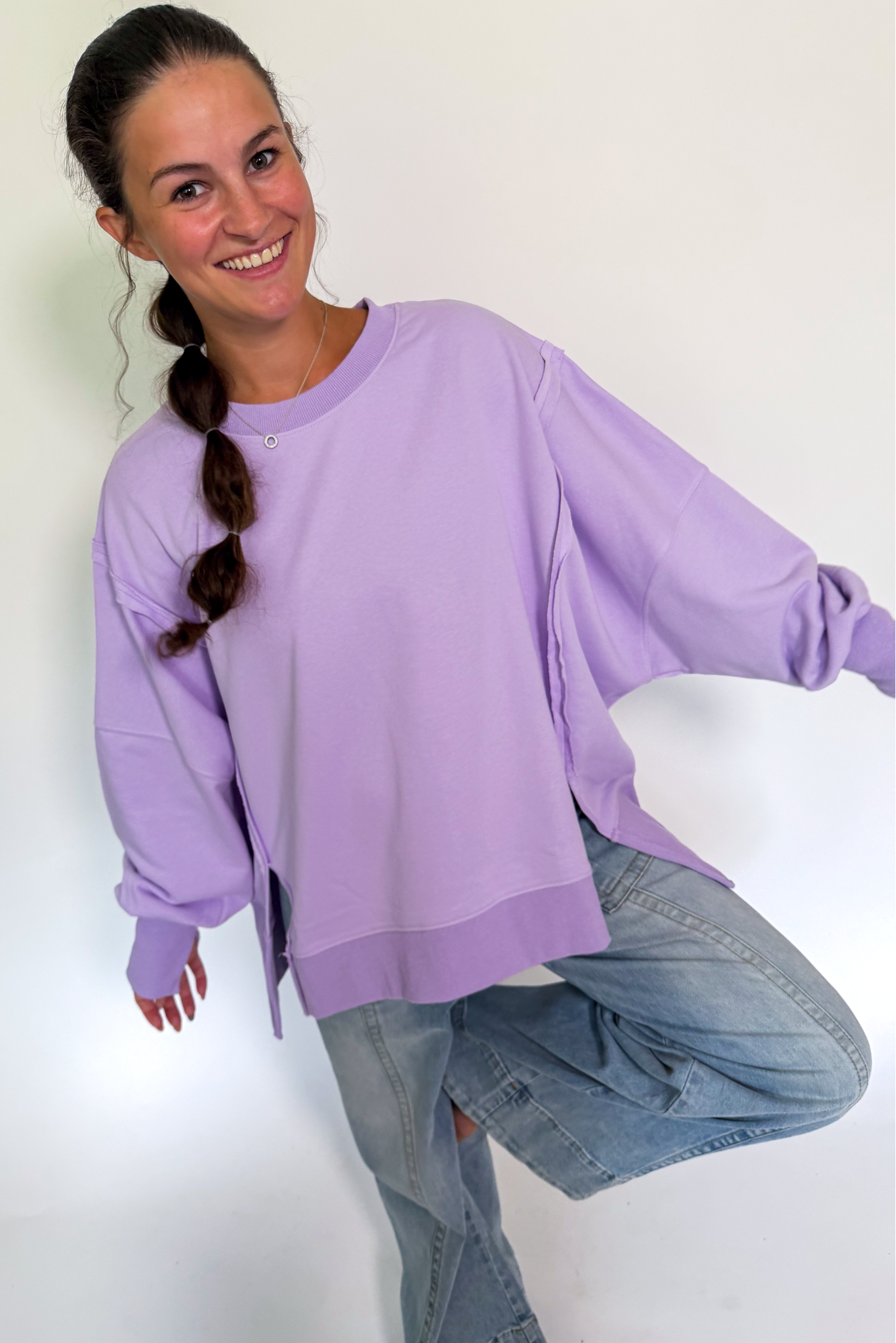 A woman wearing an oversized light purple sweatshirt and light blue jeans, posing against a plain white background.