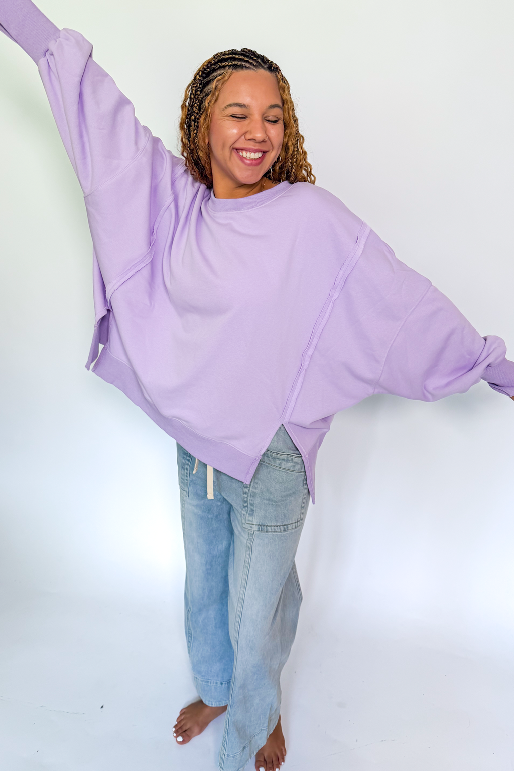 A woman modeling an oversized lavender-purple long-sleeve sweatshirt with wide batwing sleeves, paired with light-wash blue jeans, against a plain white studio background.