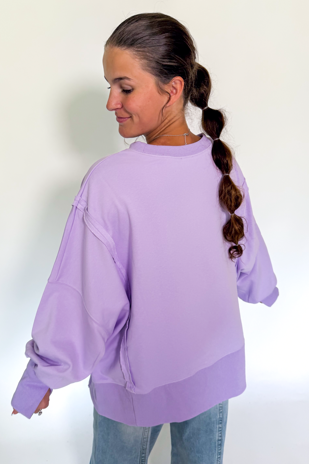 A woman with a long brown braid is shown from the back wearing an oversized light purple sweatshirt and blue jeans against a plain white background.
