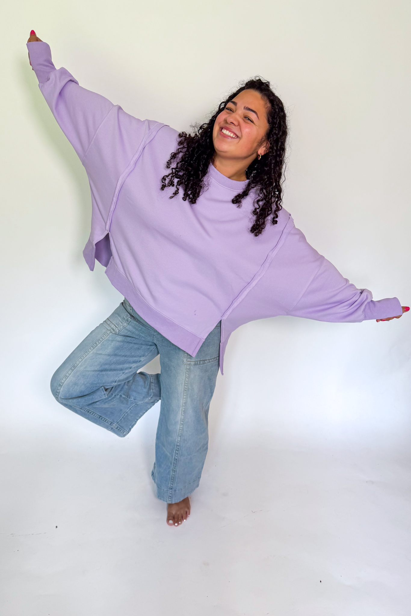 A woman posing in a studio setting wearing a lavender oversized batwing sweatshirt and light blue jeans, balancing on one leg with her arms outstretched against a plain white background.