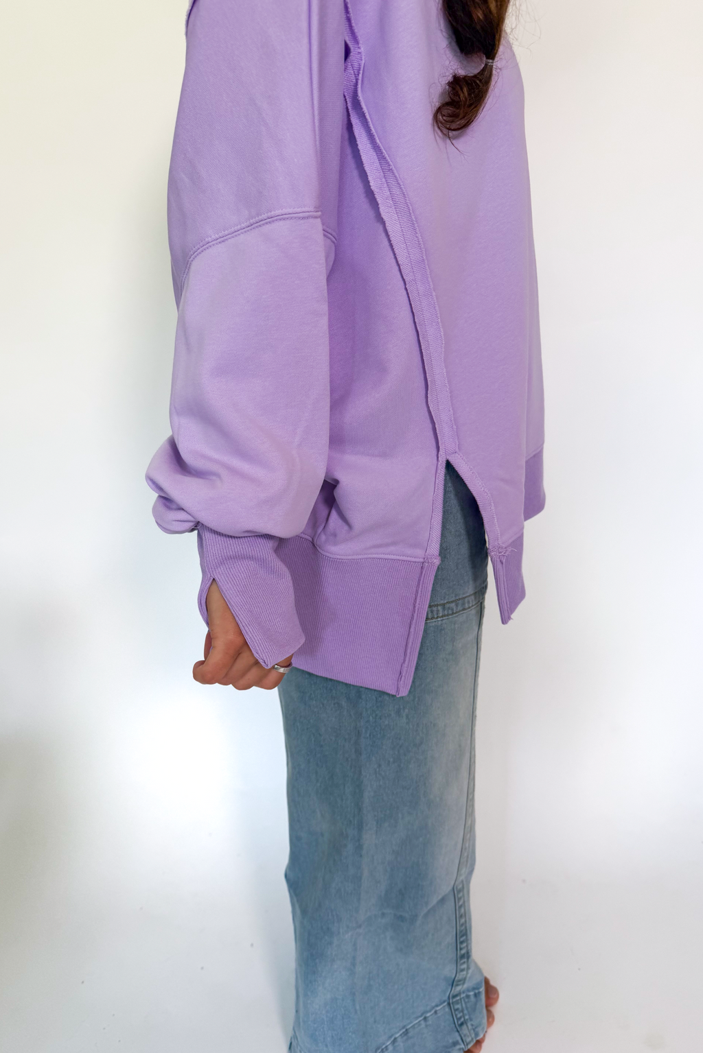 A person wearing an oversized lavender sweatshirt with a side slit and long sleeves, paired with light blue denim jeans, shown from the side against a plain white background.