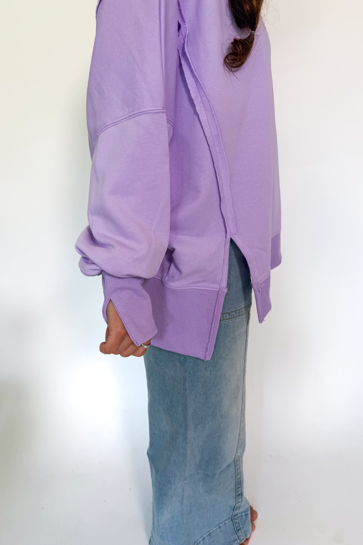 A person wearing an oversized lavender sweatshirt with a side slit and long sleeves, paired with light blue denim jeans, shown from the side against a plain white background.