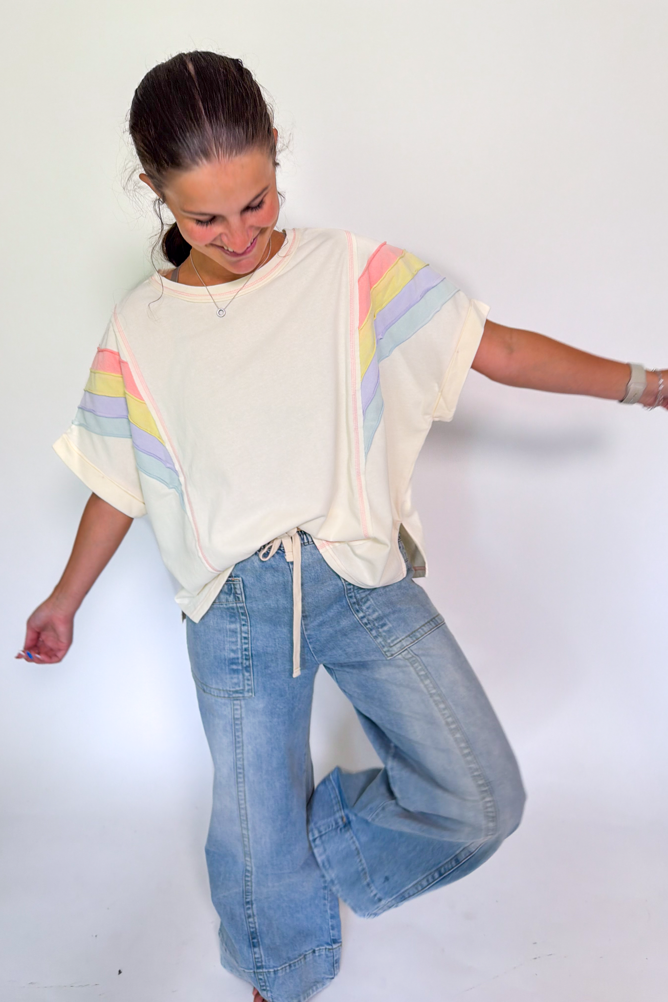 A woman modeling a cream-colored short-sleeve t-shirt with pastel rainbow stripes on the sleeves, paired with wide-leg light blue jeans, against a plain white studio background.
