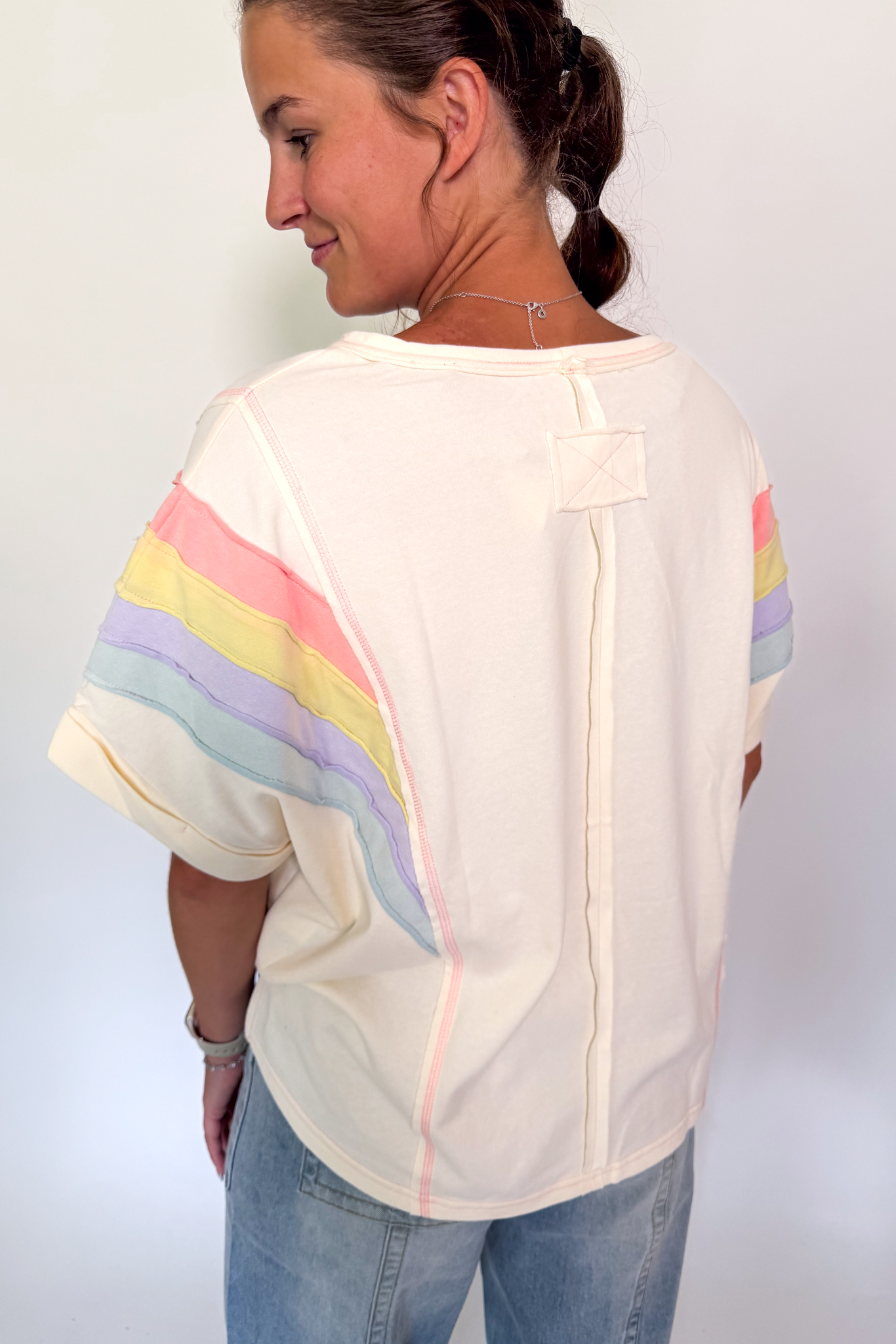 A woman shown from the back wearing a cream-colored short-sleeve t-shirt with pastel rainbow stripes on the sleeves, paired with light blue jeans.