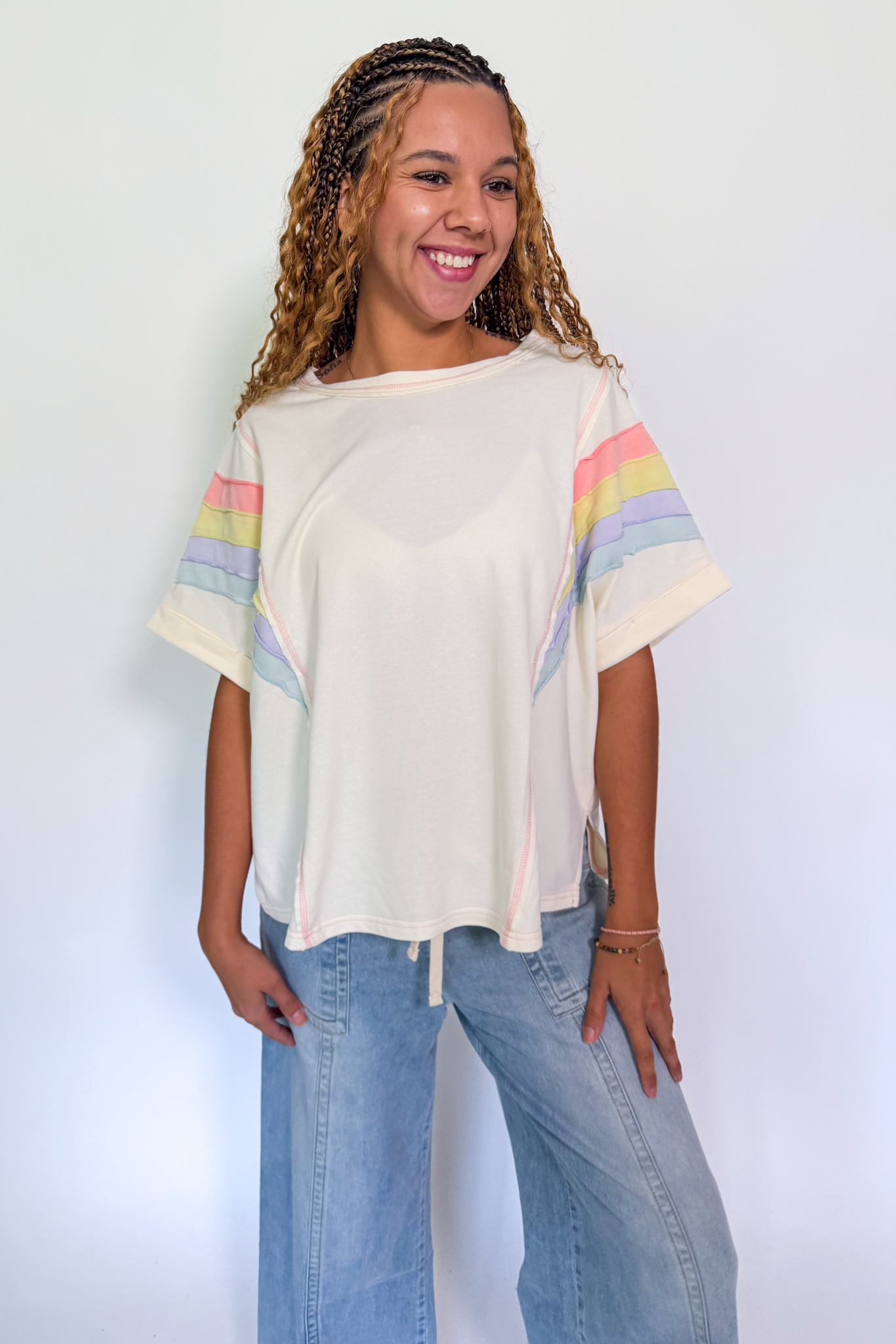 A smiling woman with braided hair wearing a cream-colored short-sleeve top with pastel rainbow stripes on the sleeves, paired with light blue jeans, standing against a plain light gray background.