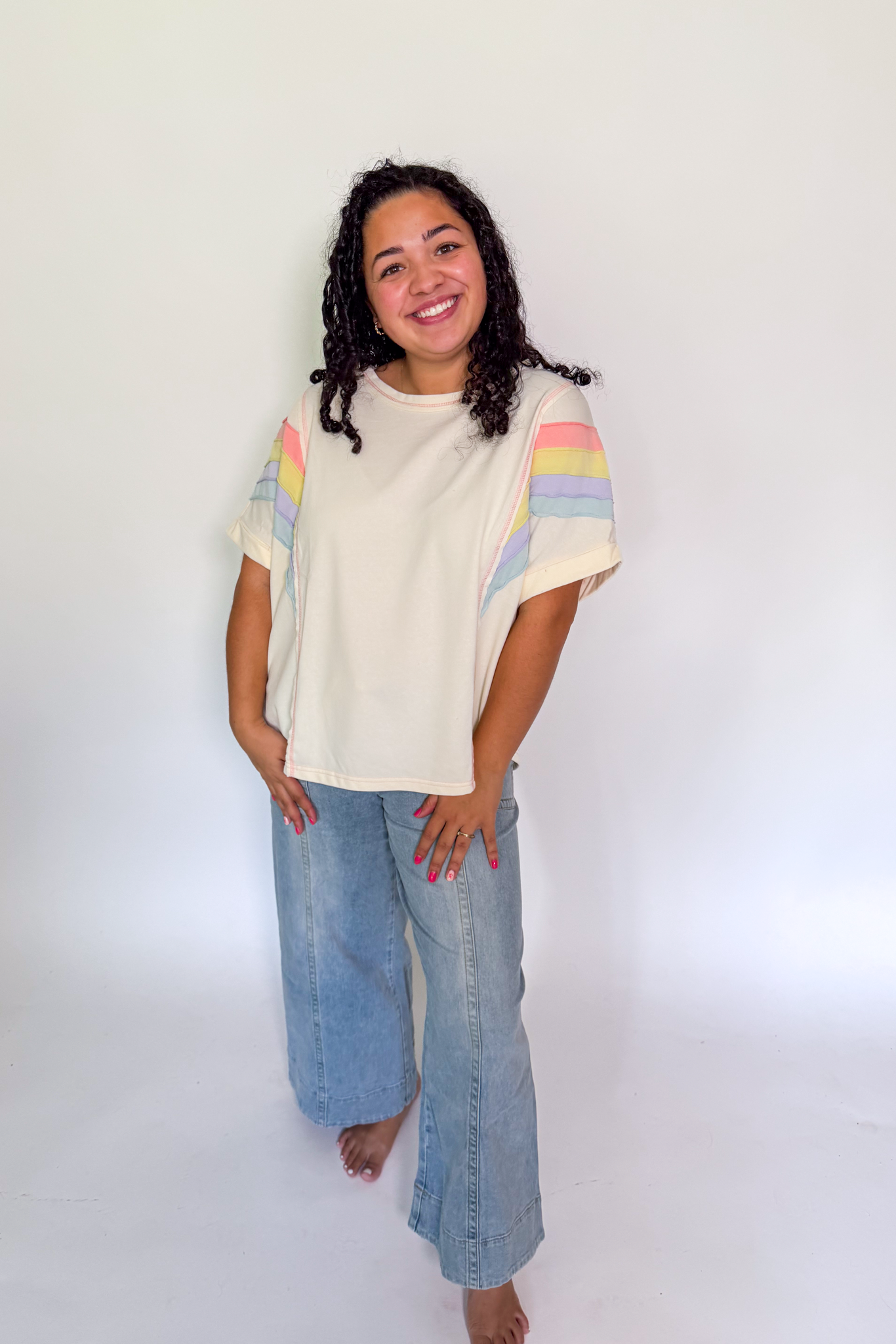 A woman modeling a cream-colored short-sleeve t-shirt with pastel rainbow stripes on the sleeves, paired with light-wash wide-leg jeans, standing against a plain white studio background.