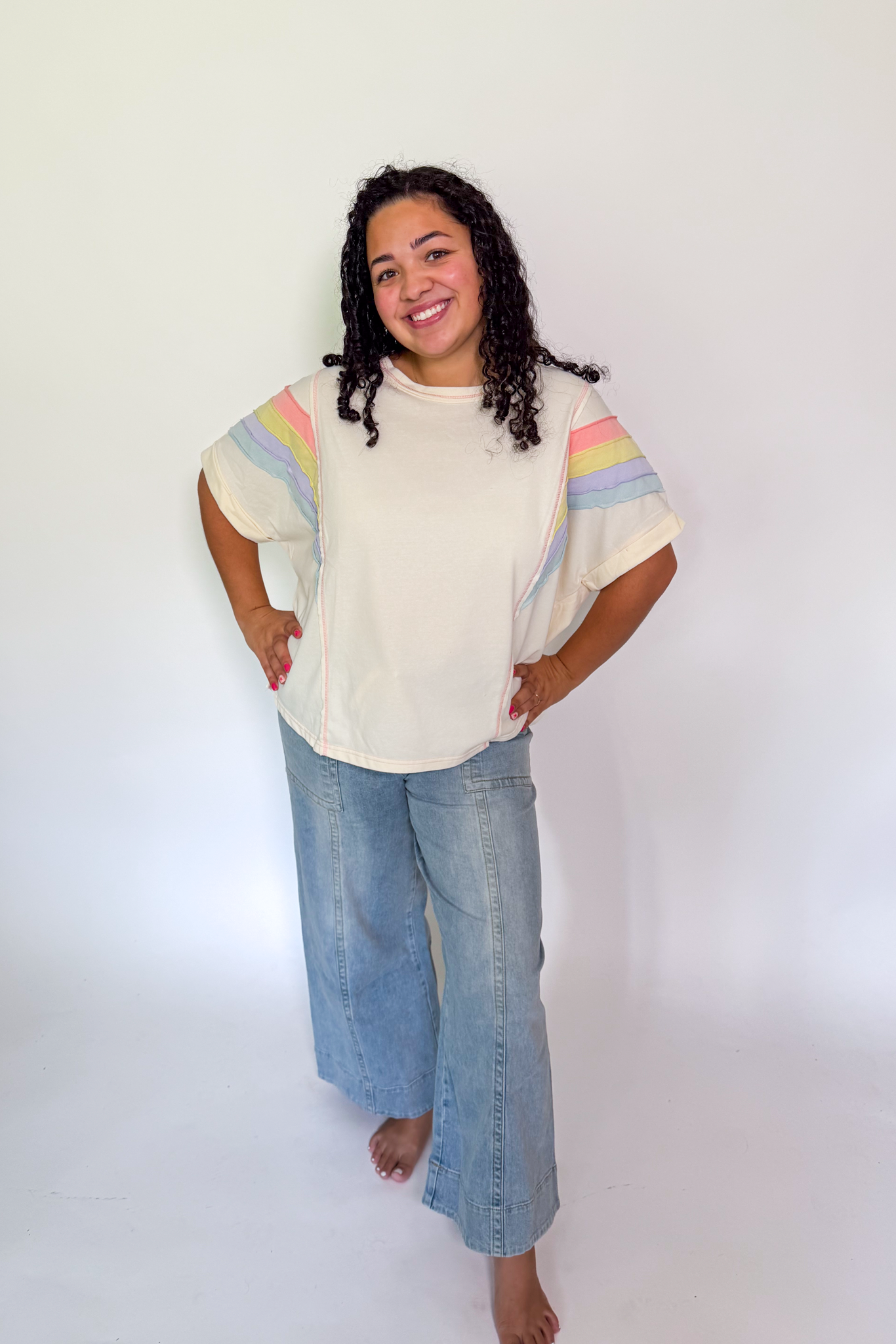 A woman modeling a cream-colored short-sleeve t-shirt with pastel rainbow stripes on the shoulders, paired with light-wash wide-leg jeans, standing against a plain white studio background.