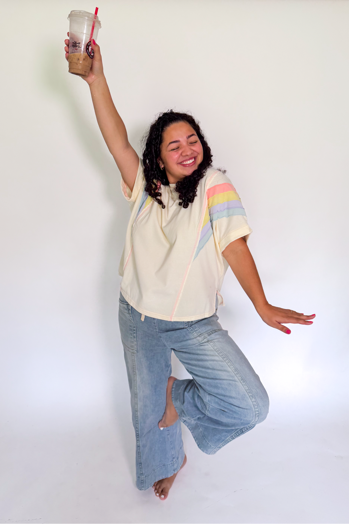 A woman posing in a studio wearing a cream-colored short-sleeve top with pastel rainbow stripes on the sleeves and light-wash blue jeans, holding a takeaway coffee cup.