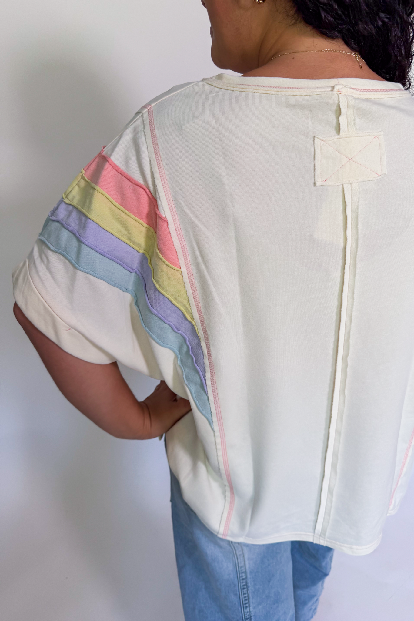 A person shown from the back wearing a cream-colored short-sleeve top with pastel rainbow stripes on the sleeves and light blue jeans, against a plain white background.