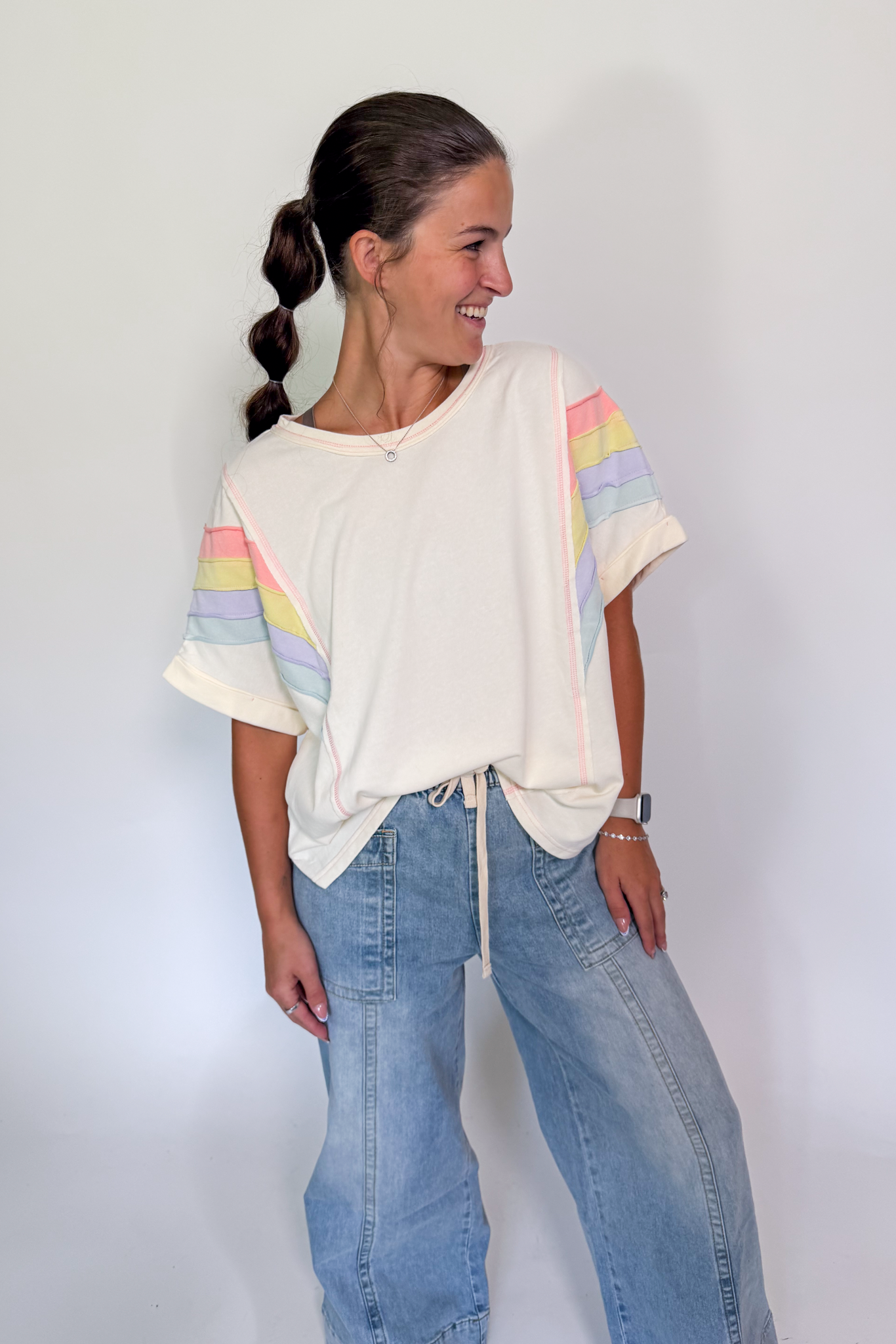 A woman wearing a cream-colored t-shirt with pastel rainbow stripes on the sleeves, paired with light-wash blue jeans, standing against a plain white wall.