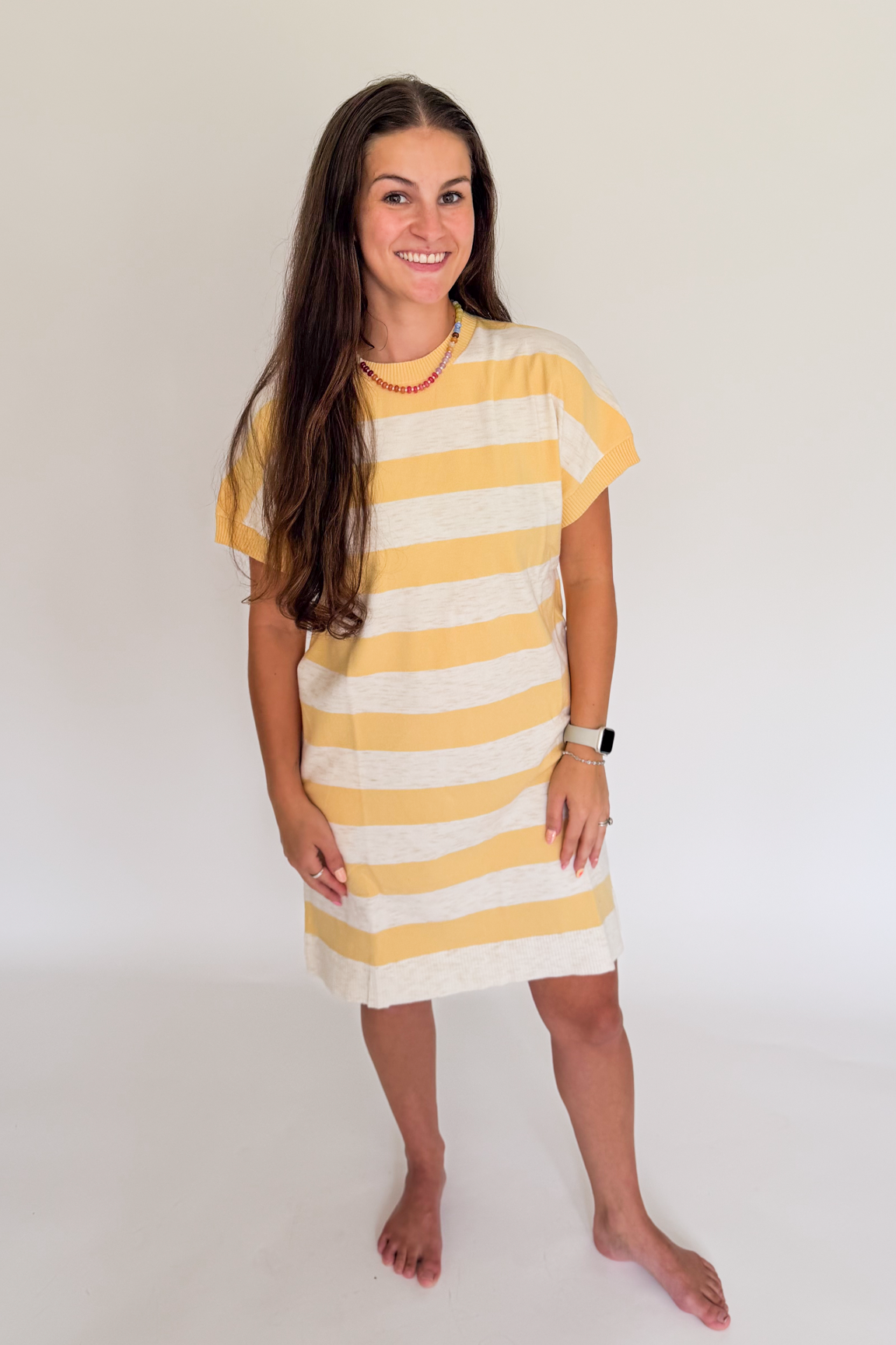 A young woman with long brown hair stands barefoot against a plain white studio background, wearing a short-sleeve, loose-fitting tunic dress with horizontal yellow and off-white stripes.