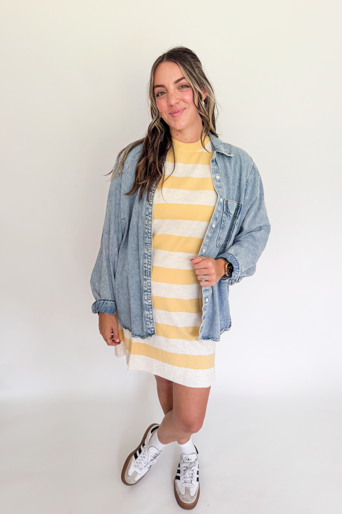 A woman modeling a yellow and white striped mock-neck dress, layered with a light blue denim shirt jacket and white sneakers, standing against a plain white studio background.