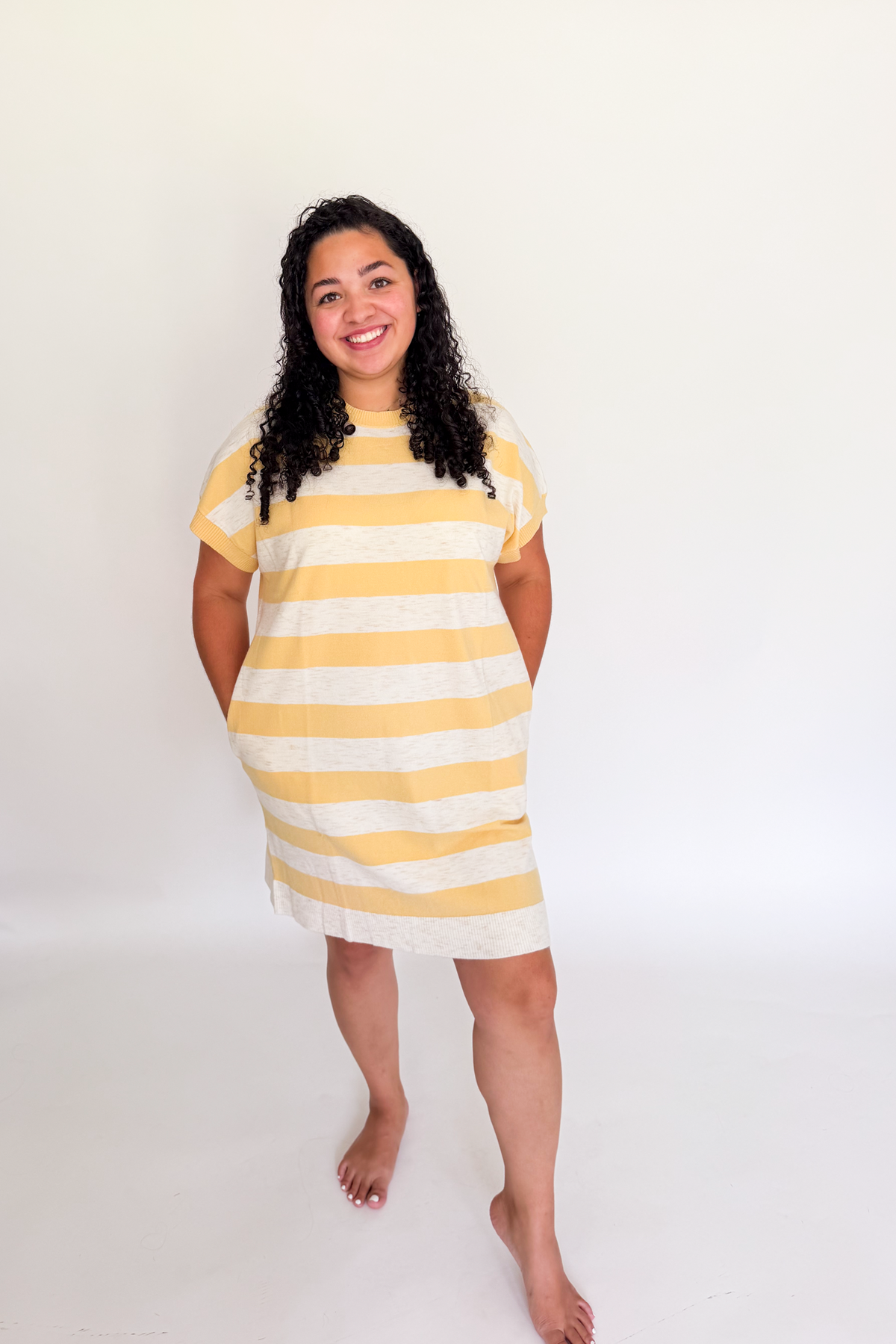 A woman modeling a short-sleeve, loose-fitting linen dress with horizontal yellow and off-white stripes, standing against a plain white studio background.
