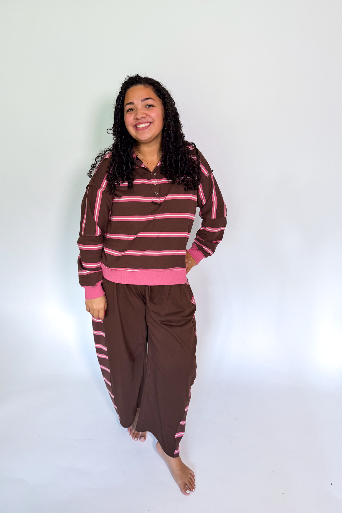 A woman modeling a two-piece lounge set consisting of a brown and pink striped long-sleeve top and matching brown pants with pink stripes, standing against a plain white studio background.