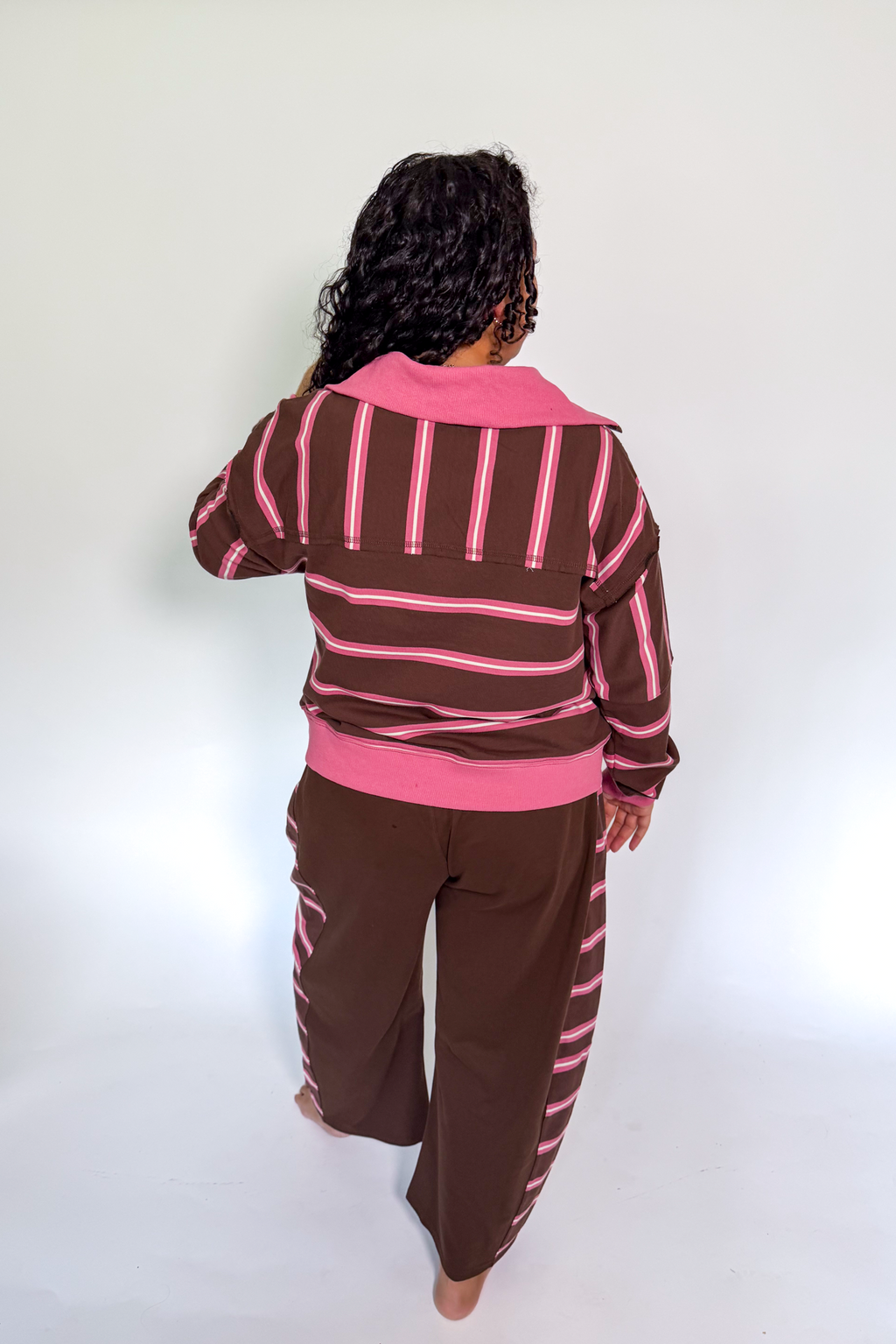 A person shown from the back wearing a brown and pink striped long-sleeve top and matching brown pants with pink stripes, standing against a plain white studio background.