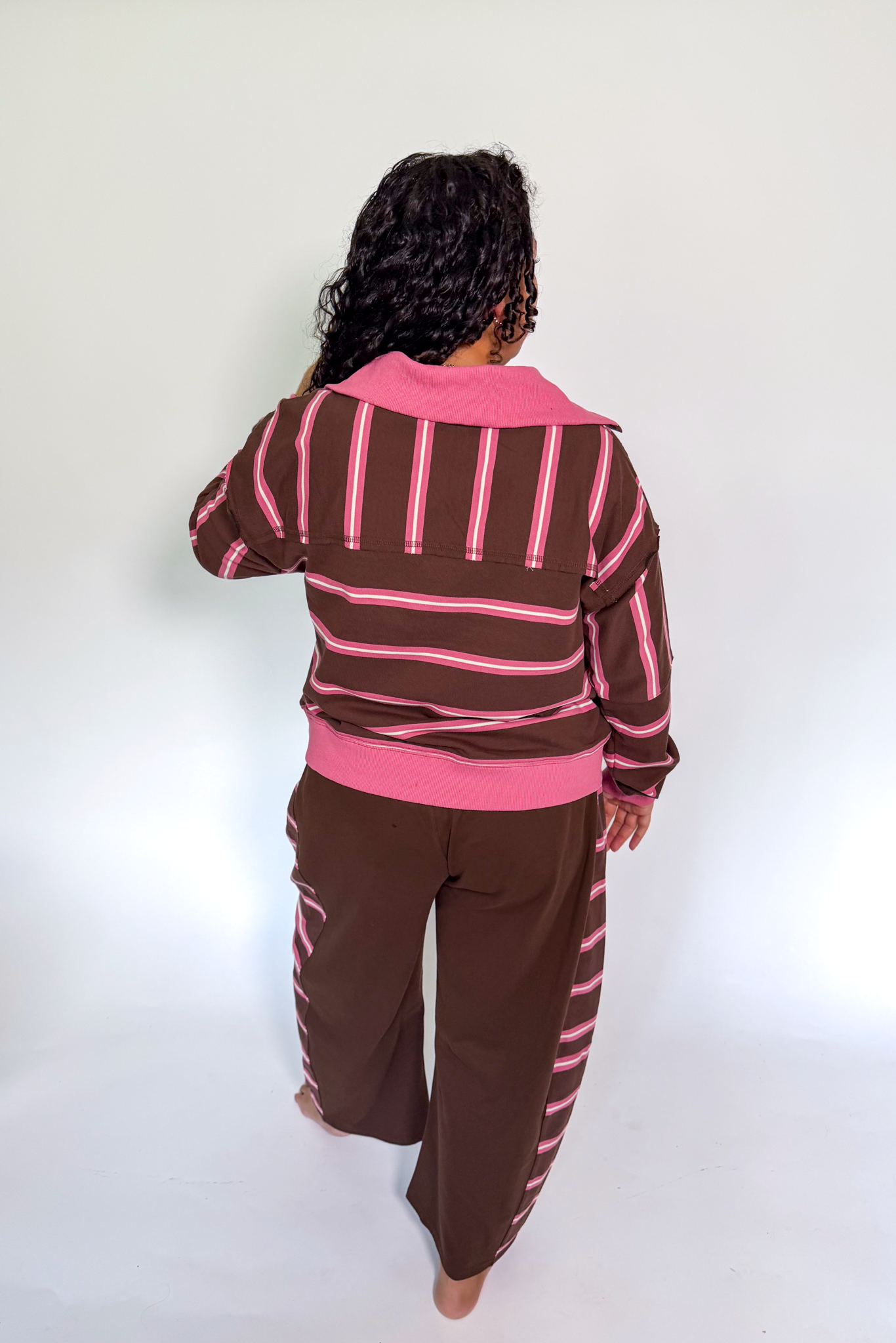 A person shown from the back wearing a brown and pink striped long-sleeve top and matching brown pants with pink stripes, standing against a plain white studio background.