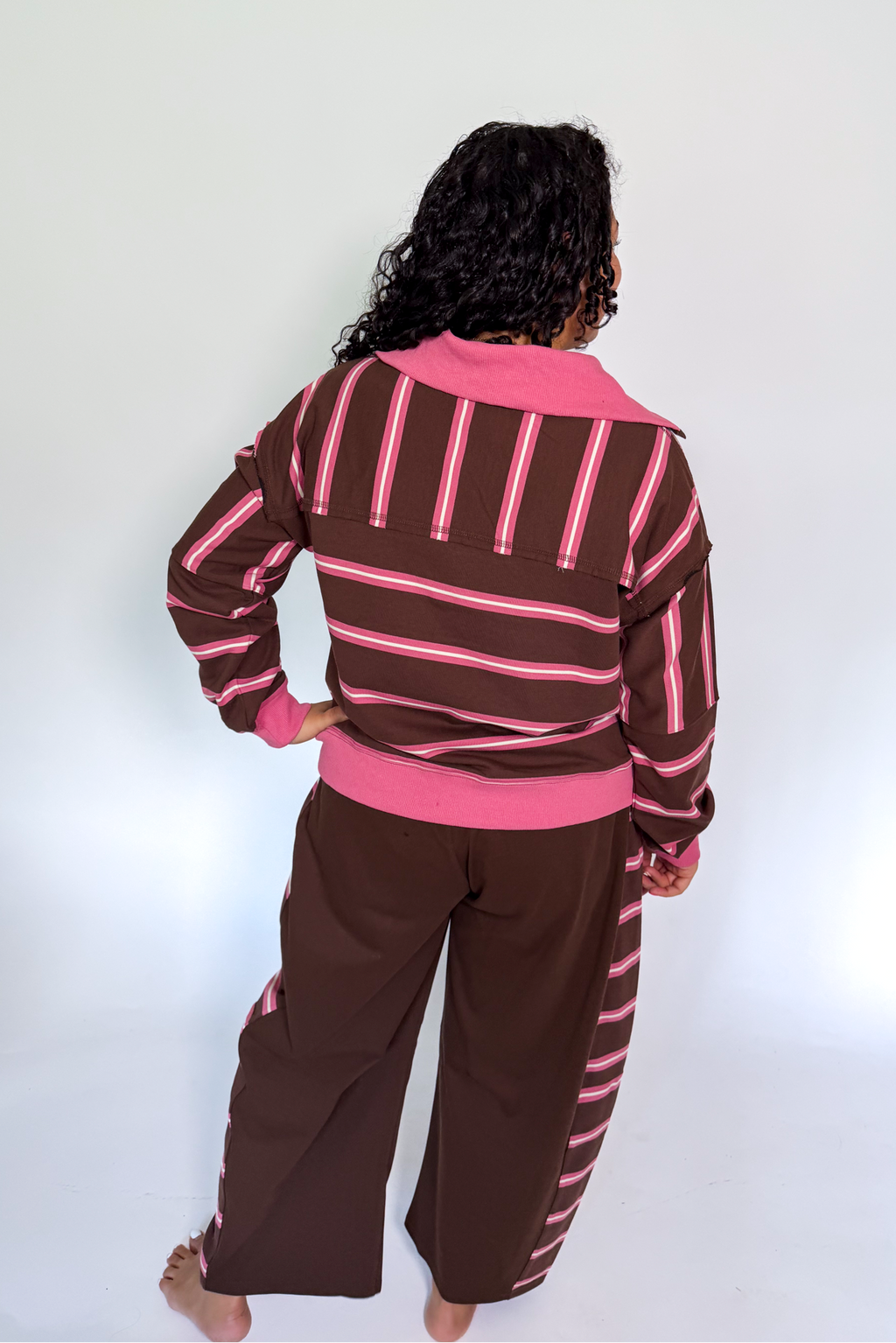 A person shown from the back wearing a brown and pink striped long-sleeve top and matching brown pants with pink stripes, standing against a plain white studio background.