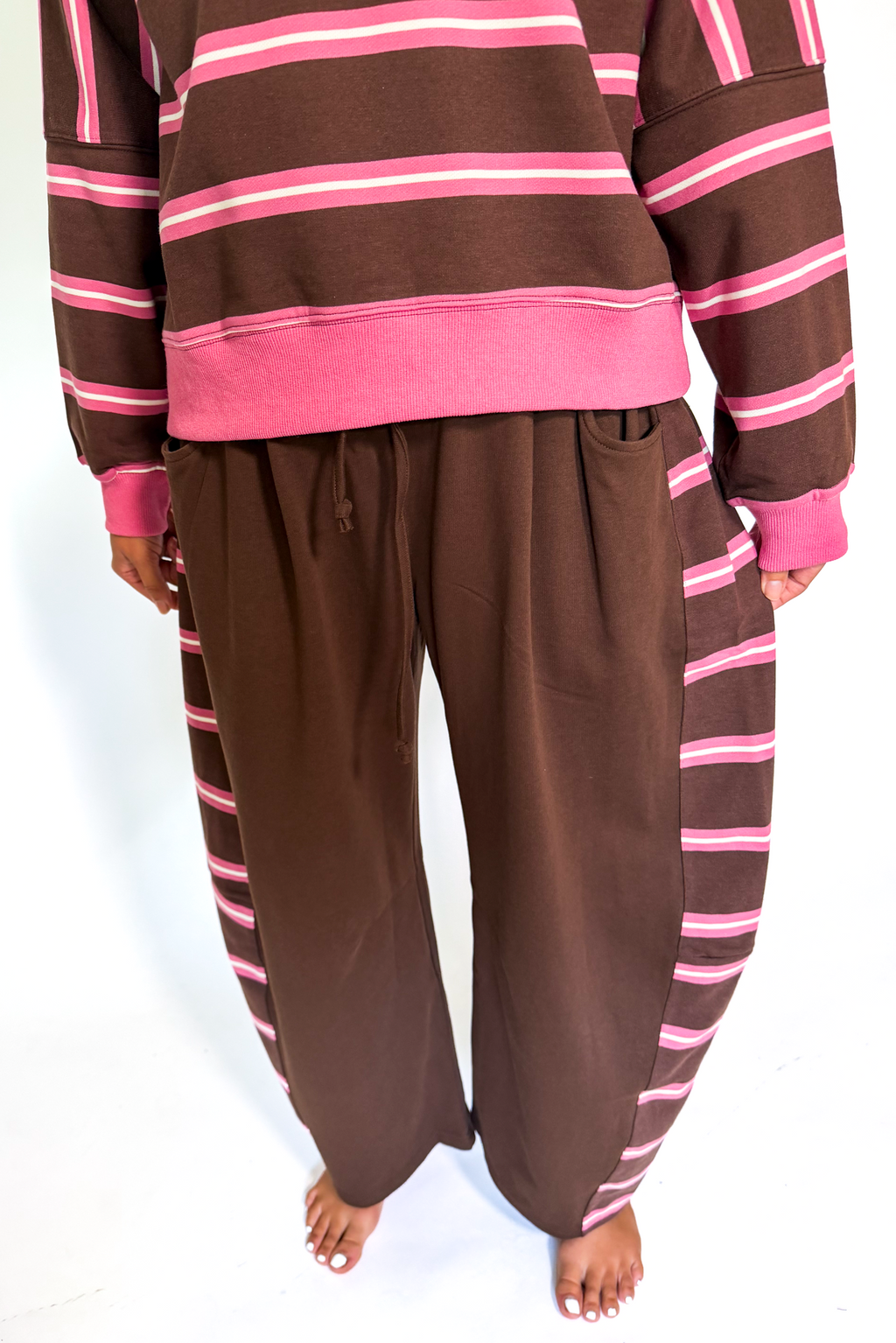 A person wearing a brown and pink striped lounge set, featuring a sweatshirt and wide-leg pants, standing against a plain white background.