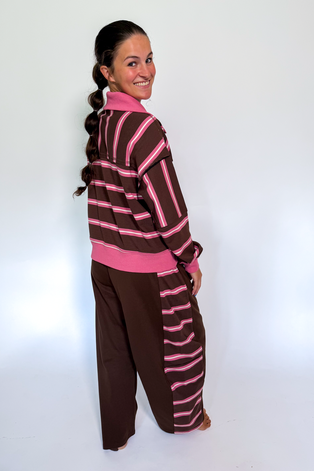 A woman modeling a brown and pink striped lounge set, featuring a long-sleeve top with a pink collar and matching wide-leg pants, photographed against a plain white studio background.