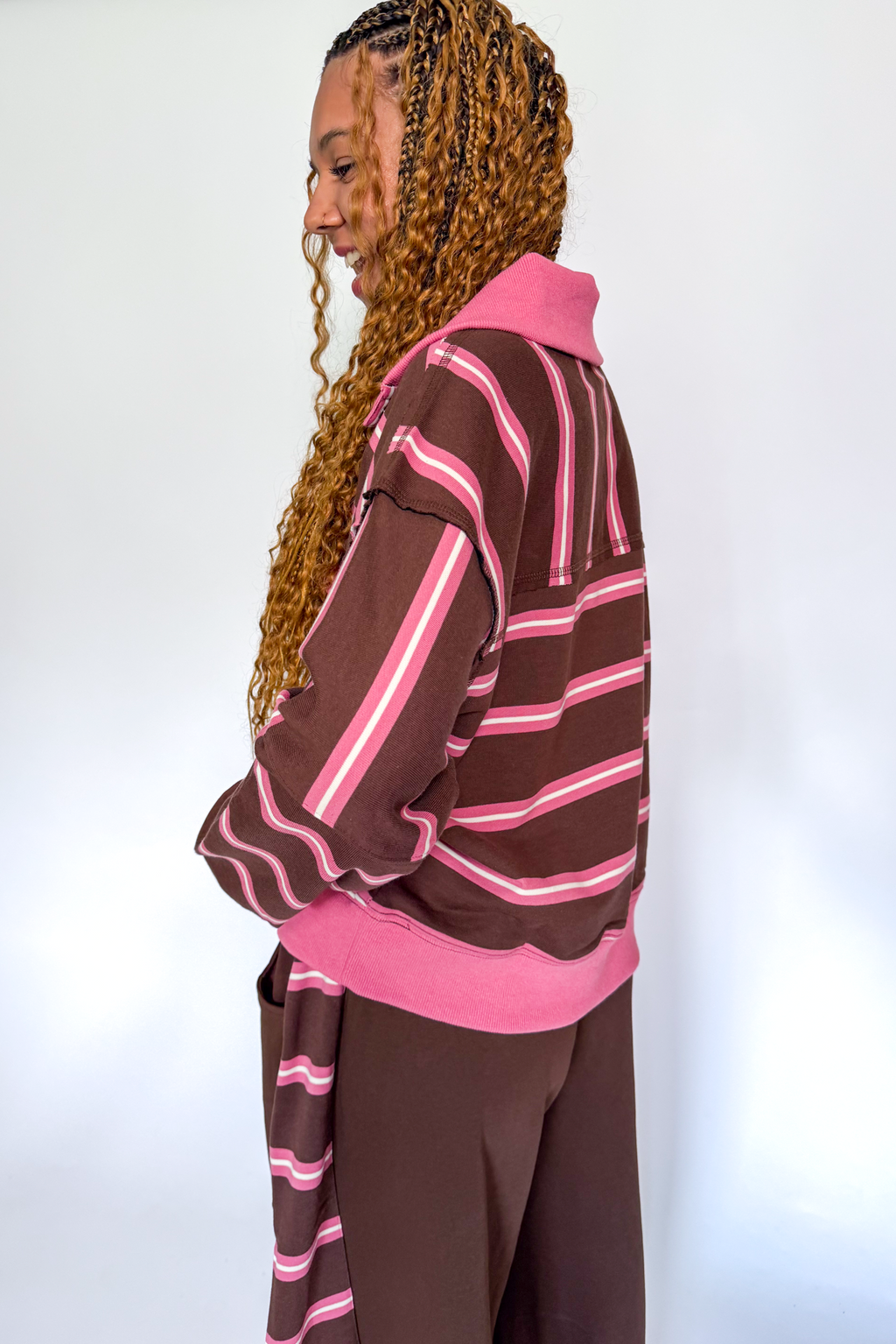 A woman with long blonde braids is shown from the side, wearing a brown and pink striped knit top and matching brown pants against a plain white background.