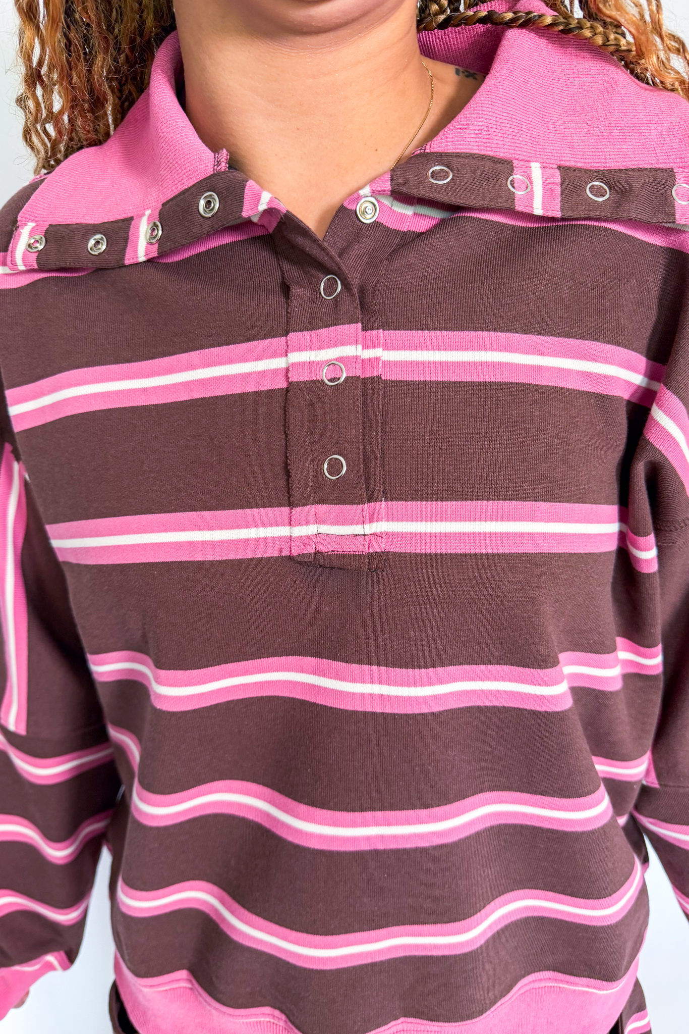 A close-up of a long-sleeve polo shirt with horizontal brown, pink, and white stripes, featuring a pink collar with snap details.