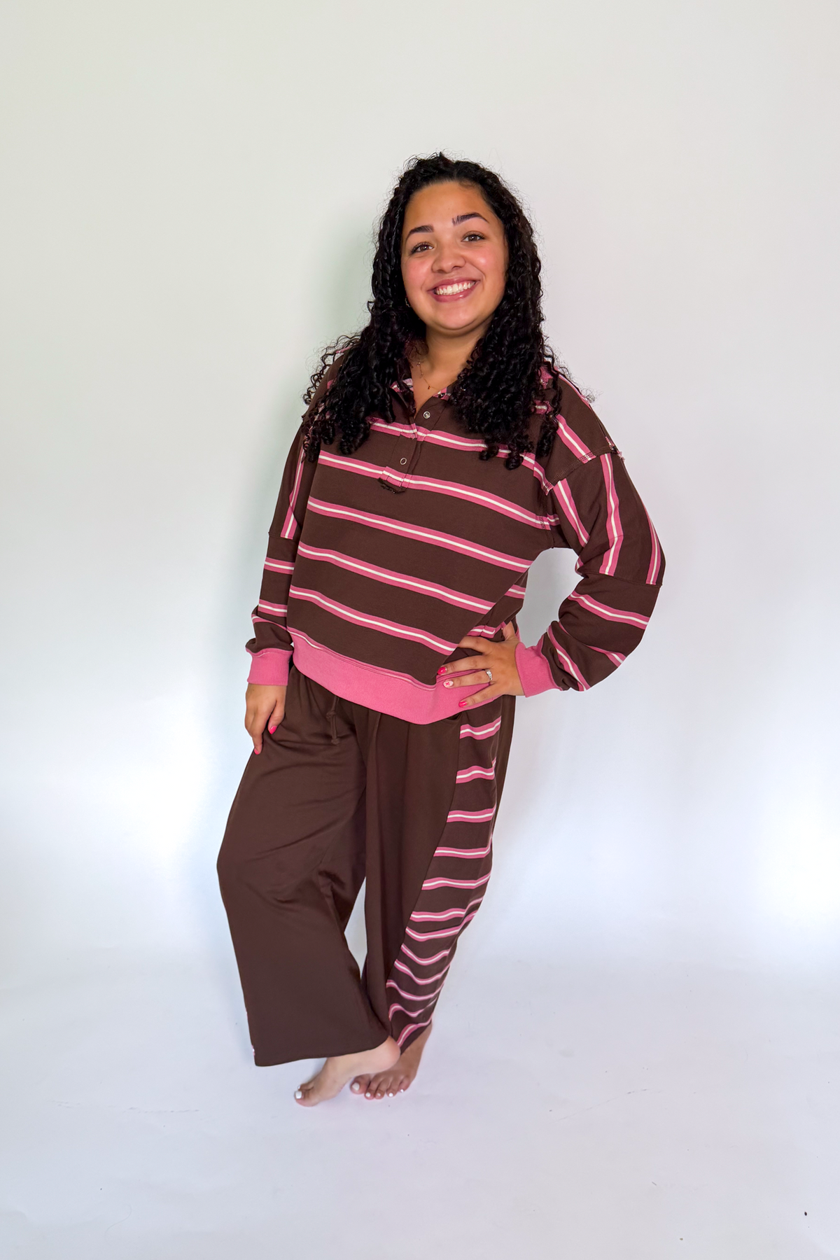 A woman modeling a matching two-piece lounge set consisting of a brown and pink striped long-sleeve top and brown pants with pink stripes, standing against a white studio background.