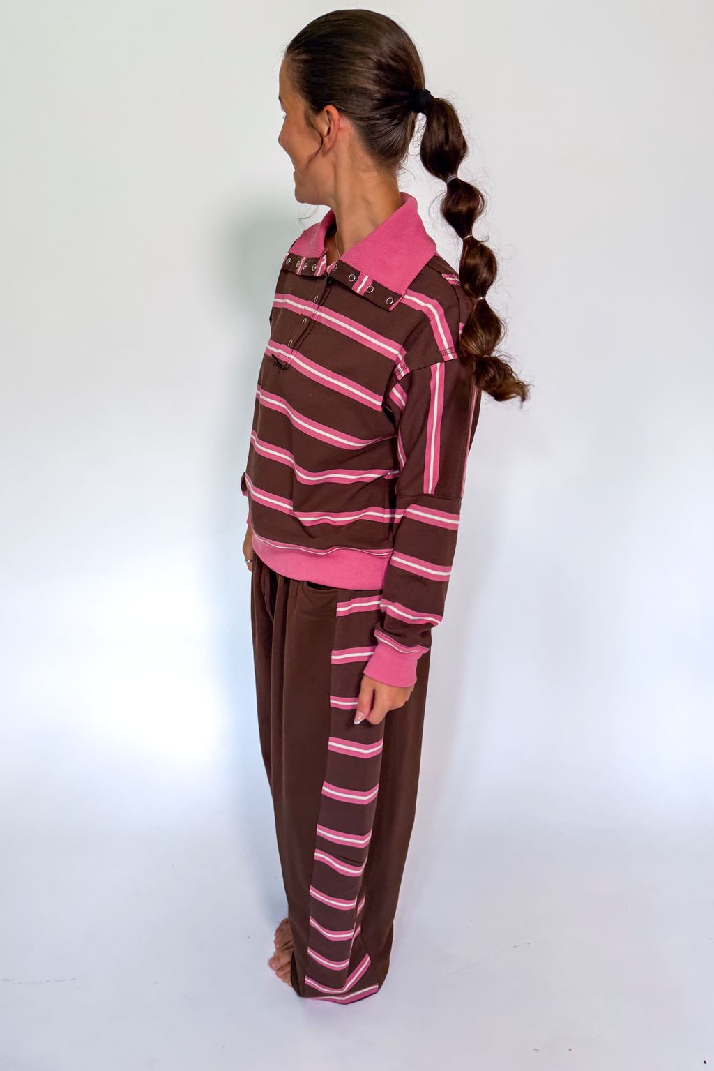 A woman stands in profile wearing a brown and pink striped long-sleeve top and matching brown pants with pink stripes, against a plain white studio background.