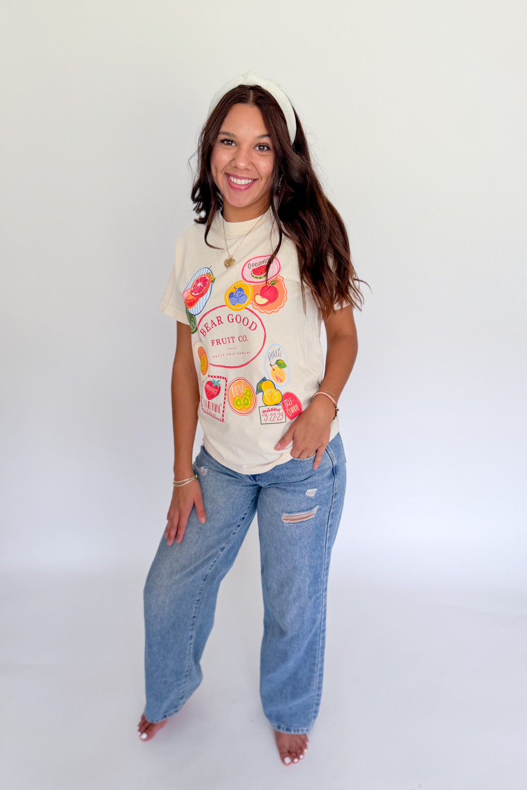 A woman modeling a cream-colored graphic t-shirt with colorful fruit illustrations and the text 'SUGAR GOOD FRUIT CO.' paired with light-wash blue jeans, standing against a plain white studio background.