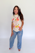 A woman modeling a cream-colored graphic t-shirt with colorful fruit illustrations and the text 'SUGAR GOOD FRUIT CO.' paired with light-wash blue jeans, standing against a plain white studio background.