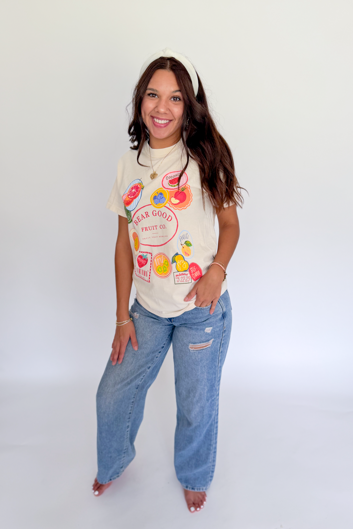 A woman modeling a cream-colored graphic t-shirt with colorful fruit illustrations and the text 'SUGAR GOOD FRUIT CO.' paired with light-wash blue jeans, standing against a plain white studio background.